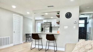 A modern living room with a sofa, two bar stools, a coffee table, and a kitchen area with a refrigerator and cabinets. - Cozy Remodeled House! (Port Richey)