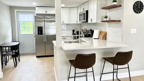 A modern kitchen with white cabinetry, stainless steel appliances, and a central island with two bar stools. - Cozy Remodeled House! (Port Richey)