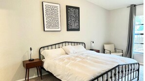A bedroom with a bed, a nightstand, a chair, and two wall art pieces.