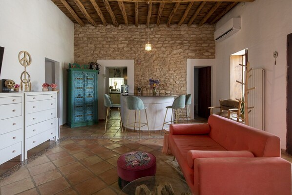 A cozy living room with a stone wall, a red sofa, a bar area with stools, and a white cabinet with drawers.