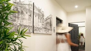 Een hal met zwart-wit kunstwerken aan de wanden, een potplant en een persoon in een witte jurk en hoed die de gang in loopt.