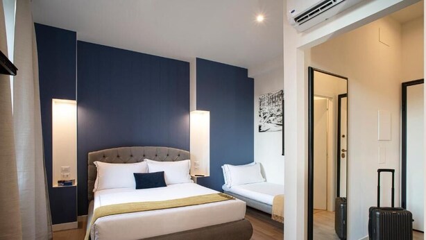 Een hotelkamer met een groot bed, een nachtkastje, een lamp, een witte bank, een spiegel en een koffer.