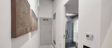 A hallway with a white door marked 'Fire Exit,' a shower area with a glass door, and a thermostat on the wall.