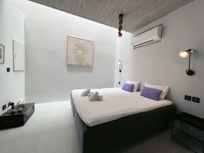 A modern bedroom with a large bed, a wall-mounted air conditioner, a framed artwork, and a bedside lamp.