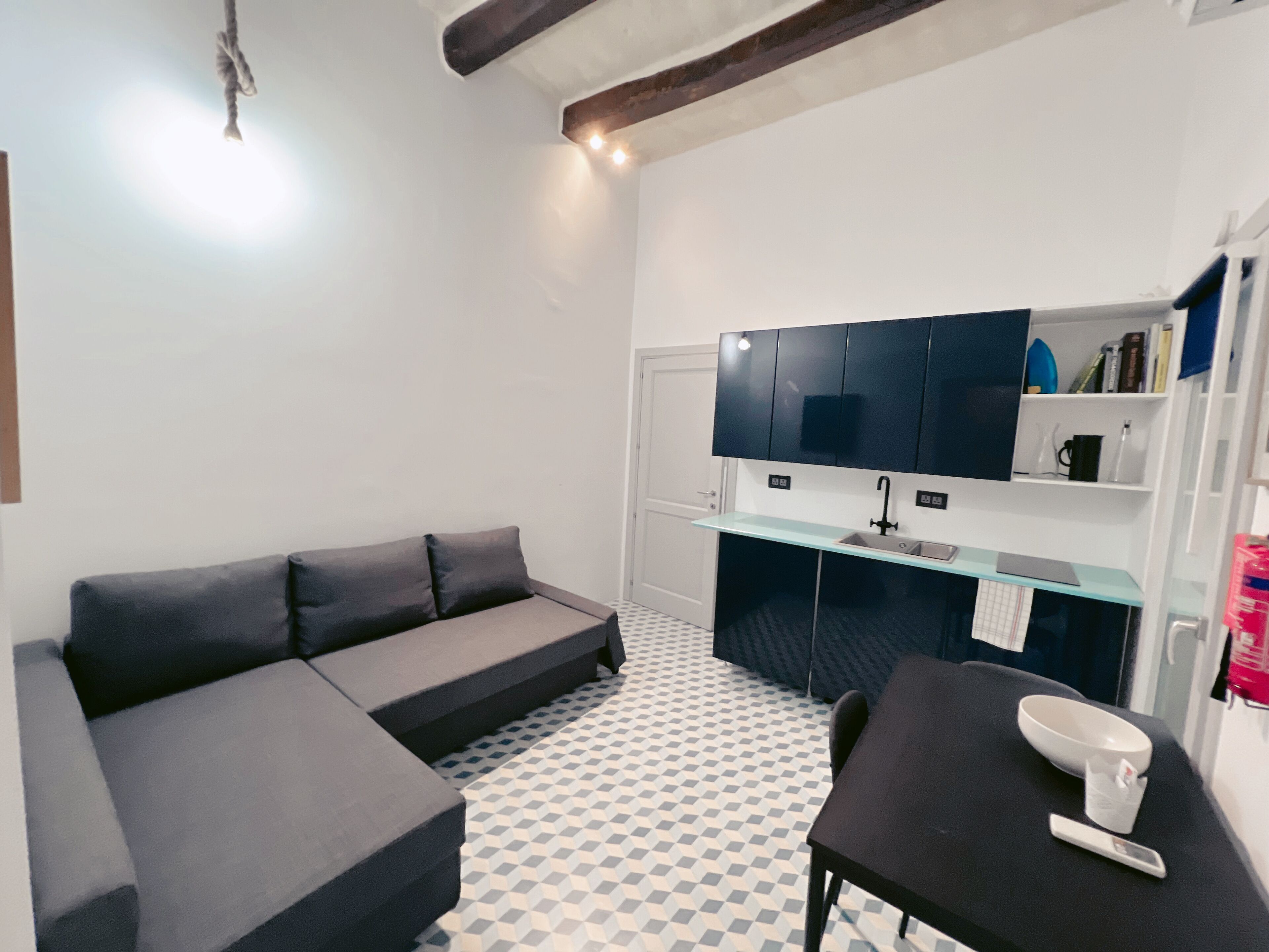 A modern, compact living space with a sofa, a small dining table, a kitchen area with cabinets, and a patterned floor.
