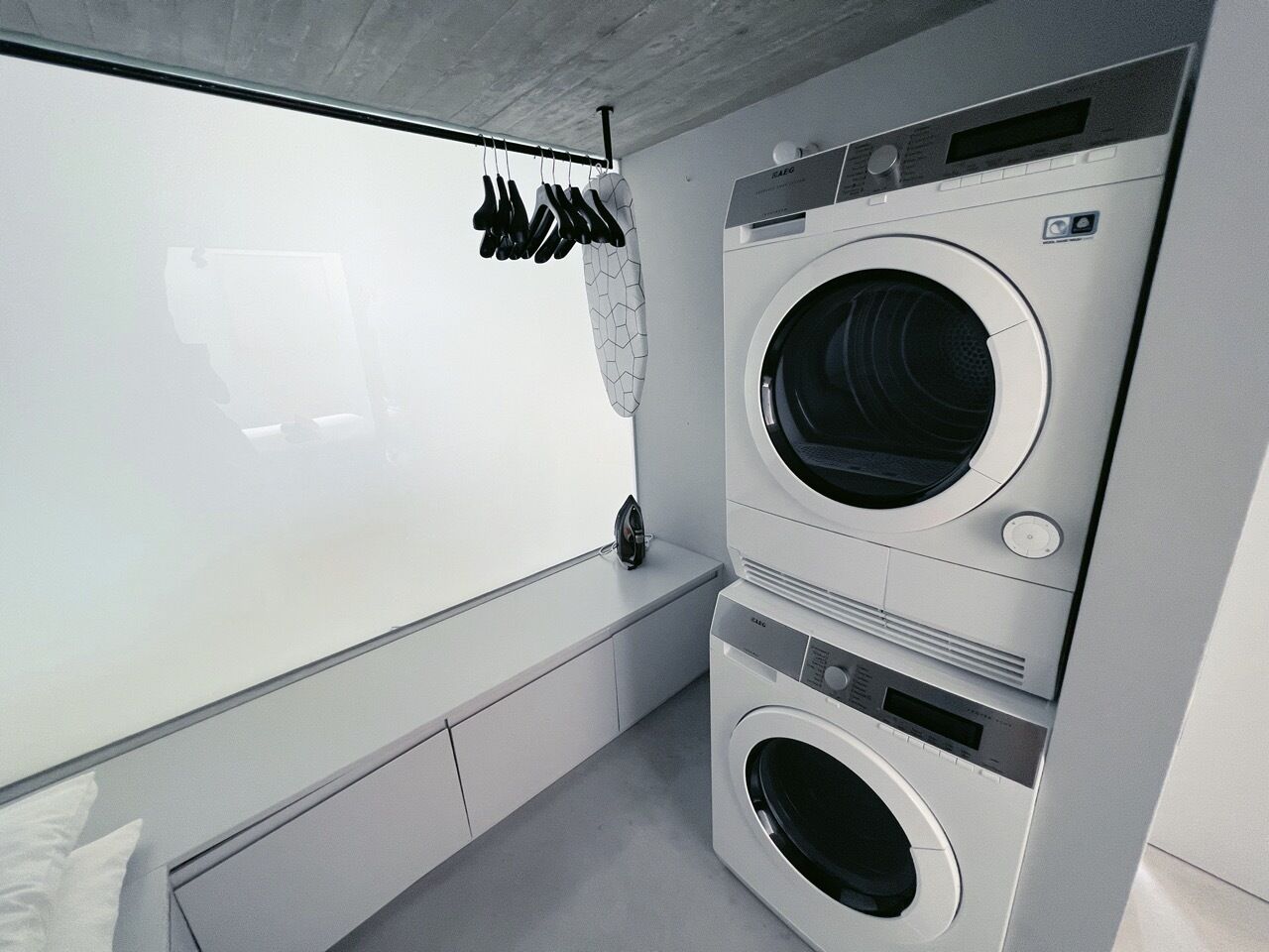 A compact laundry room with a front-loading washer and dryer, a drying rack, and a window for natural light.