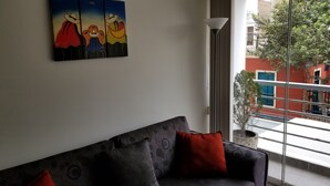 Un salon avec un canapé foncé, des coussins rouges et une grande fenêtre donnant sur un bâtiment rouge et un arbre.