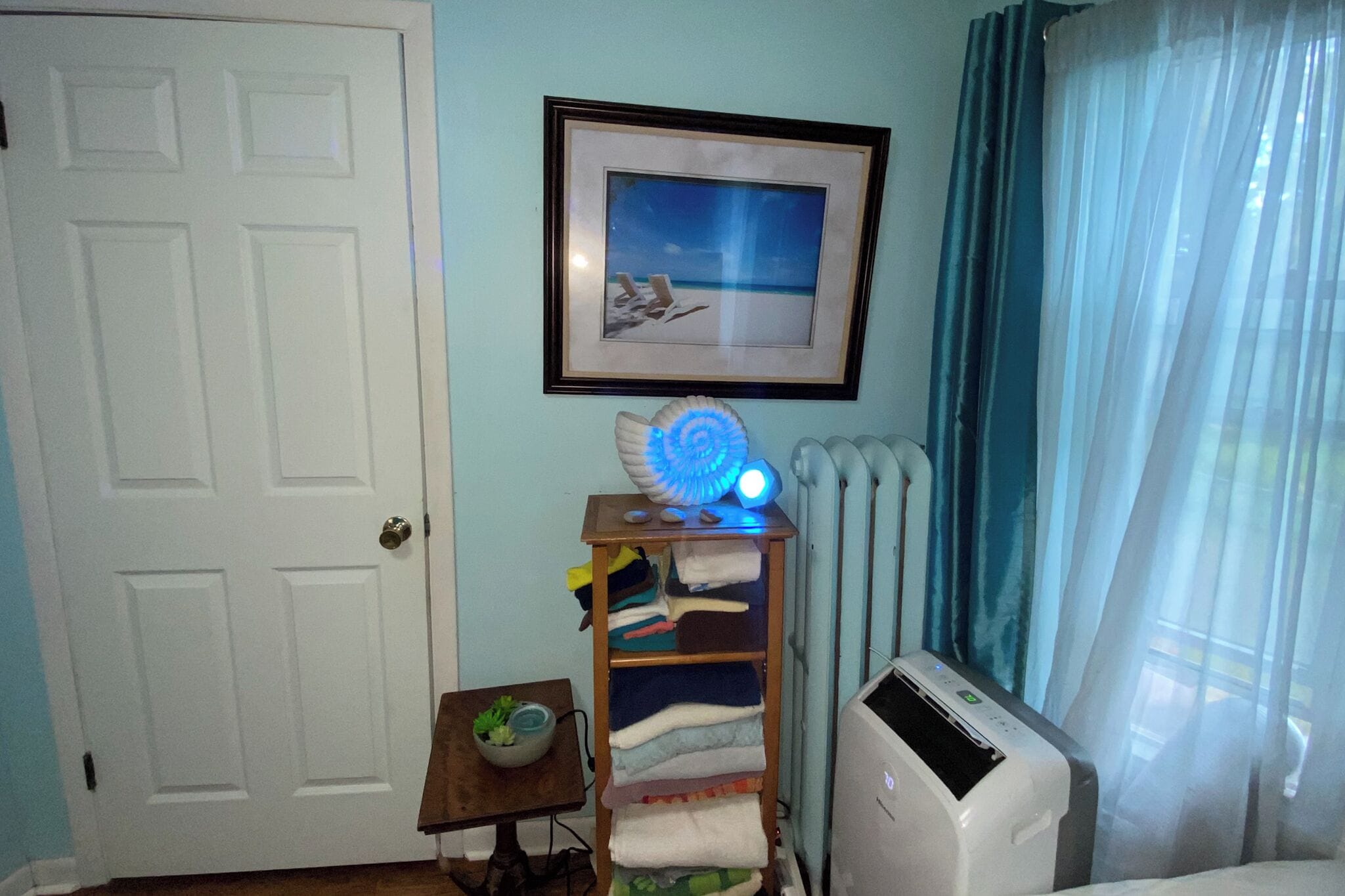 Una habitación con una puerta blanca, un cuadro enmarcado con una escena de playa, un radiador, un estante con ropa doblada y una mesita con una planta.