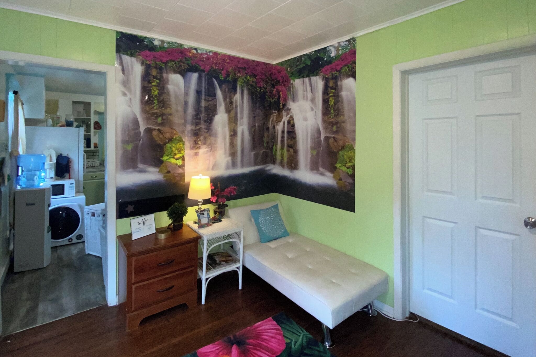 Ein Zimmer mit einem Wandgemälde eines Wasserfalls, einer weißen Bank, einem kleinen Tisch mit einer Lampe und einer Tür.