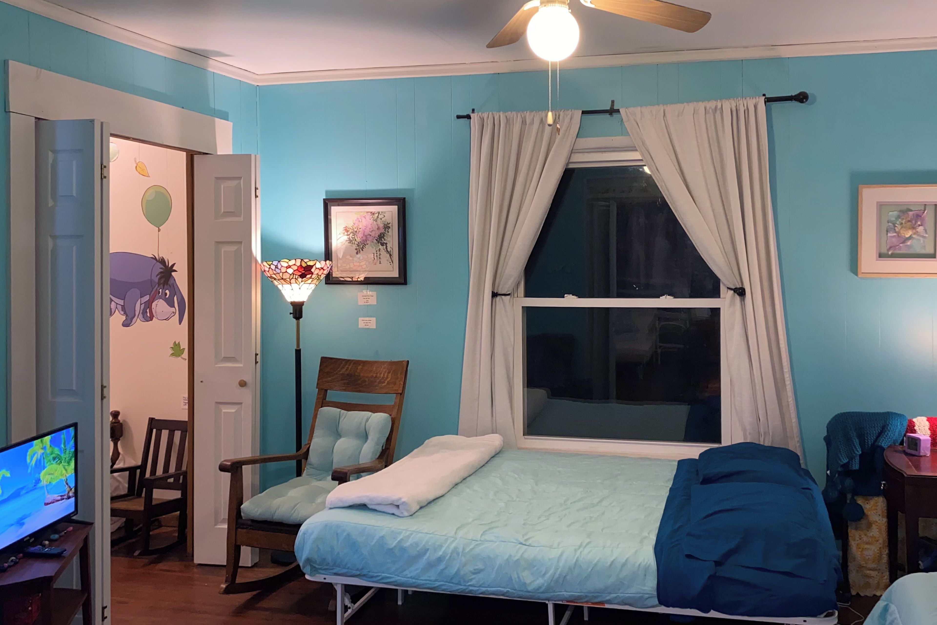 A bedroom with a bed, a chair, a desk with a computer, a window with curtains, and a ceiling fan.