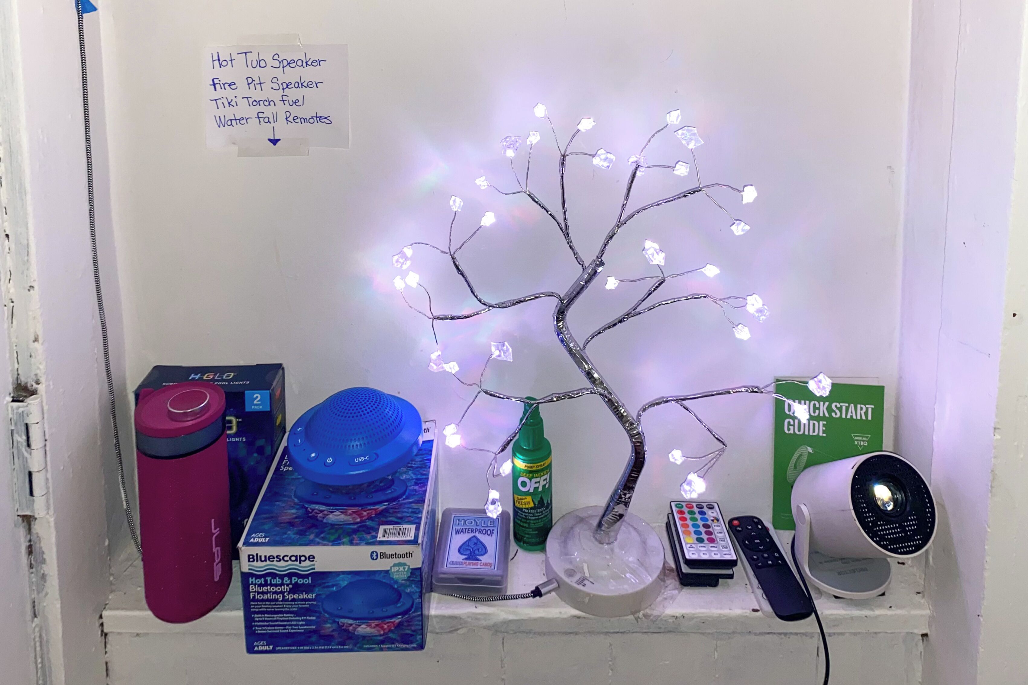 Une étagère contenant divers objets, notamment un haut-parleur Bluetooth, une bouteille d’eau, une télécommande et une guirlande lumineuse décorative en forme d’arbre.