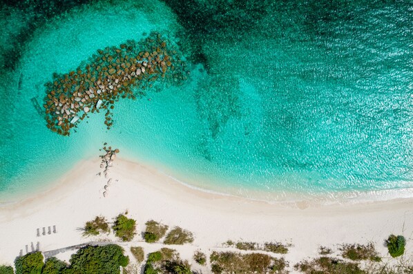Vue aérienne d’une plage aux eaux turquoise et limpides, bordée d’un rivage sablonneux et parsemée de rochers.