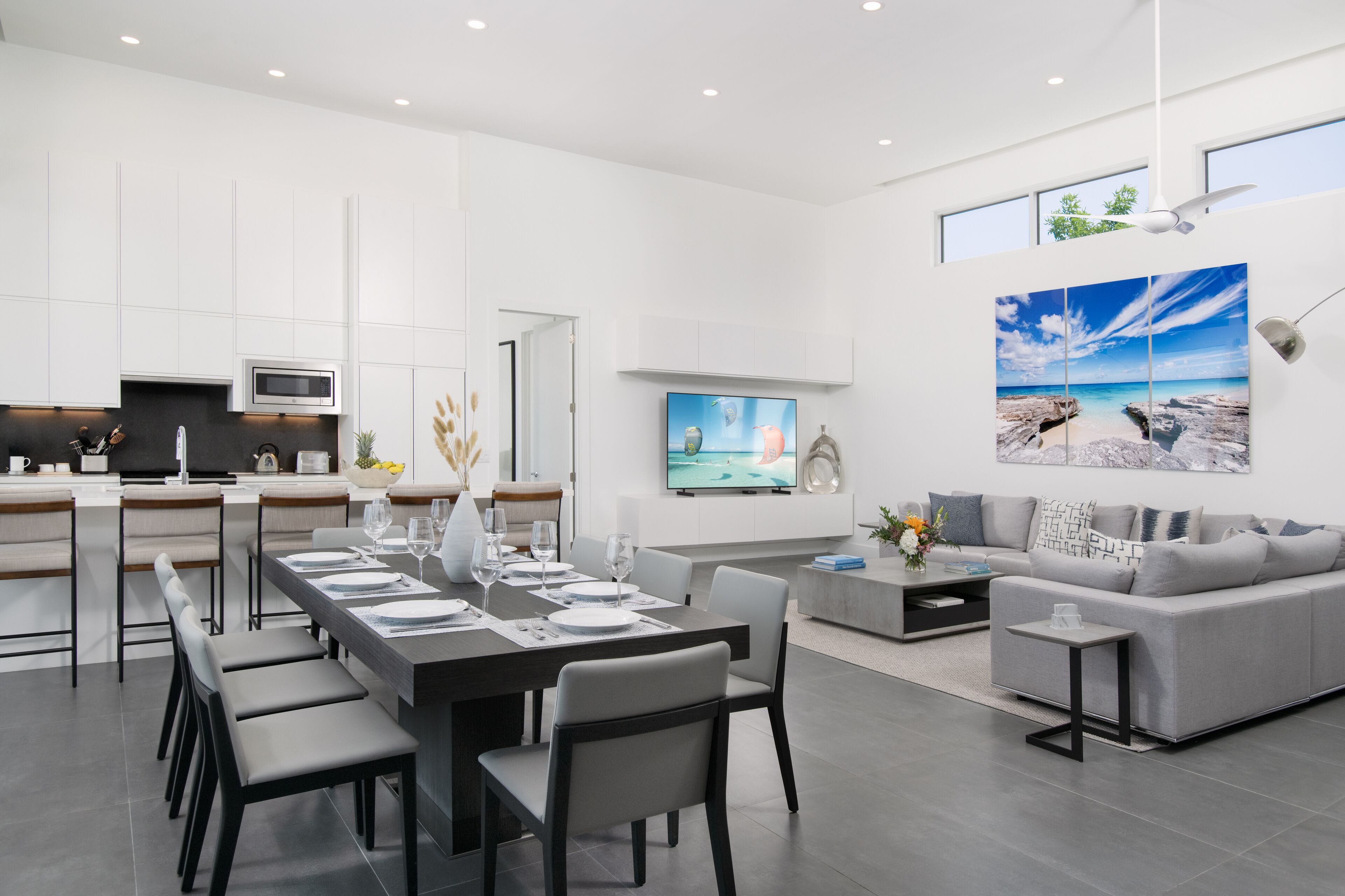 A modern living room with a grey sofa, a dining area with a long table, and a kitchen with white cabinets and a microwave.