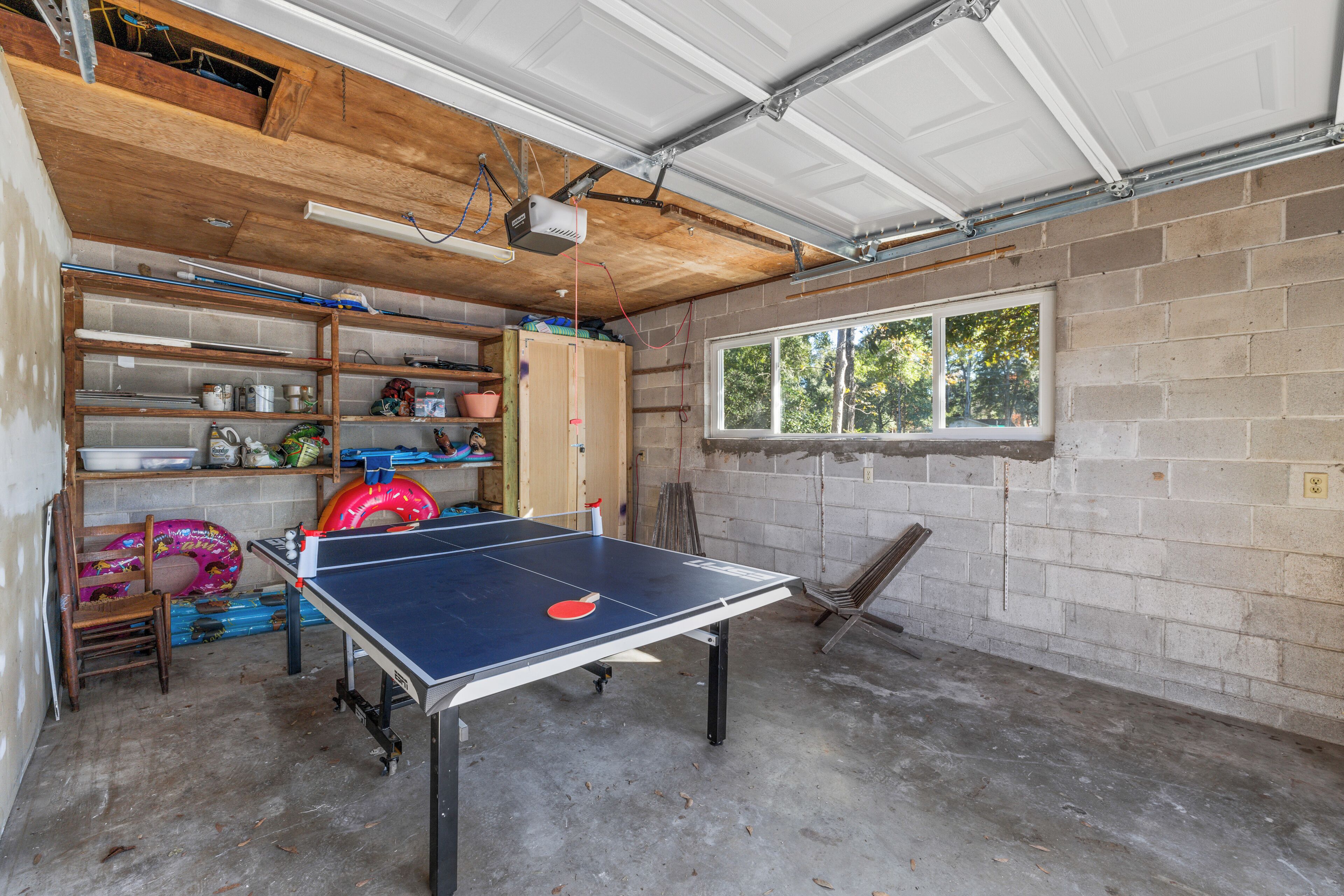 A garage with a ping pong table, shelves with various items, and a window with a view of trees.