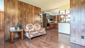 Un salon avec un plancher en bois, un canapé, une petite table avec une plante, et une cuisine en arrière-plan.