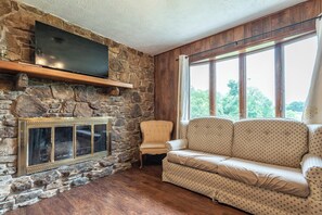 A stone fireplace with a flat-screen TV above it, a wicker chair, and a patterned sofa in a room with wooden walls and large windows. - Harmony Hills -- 3 bedroom country home (Bloomington)