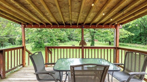 Une terrasse extérieure couverte, dotée d’une table et de chaises, entourée d’une rambarde en bois, et offrant une vue sur un paysage verdoyant.