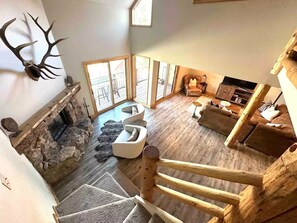 A spacious living area with a stone fireplace, a wooden staircase, and a large window overlooking a deck. - Ski in/Out! 3BR+Loft, Hot Tub/Free Parking & more! (Crested Butte)
