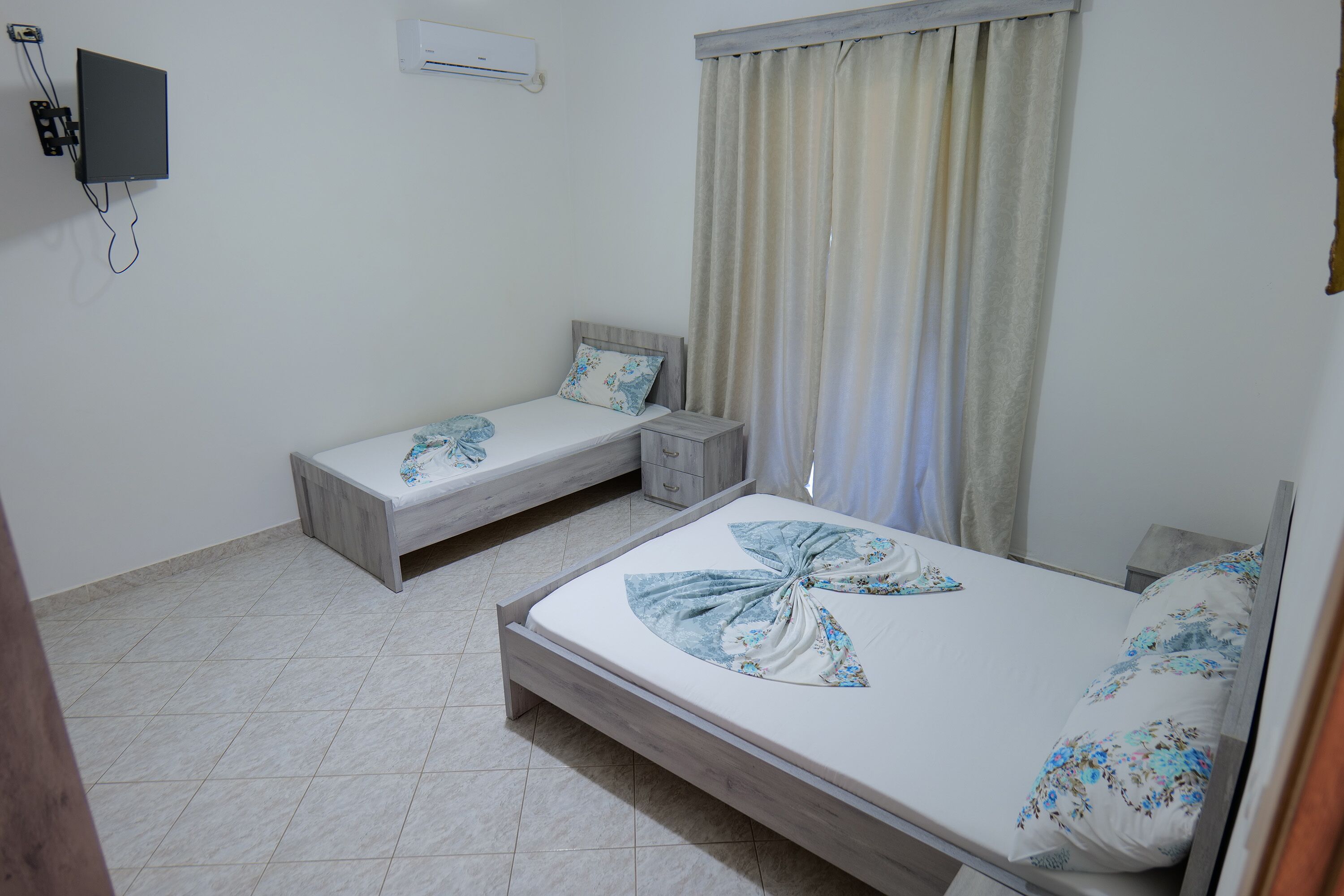 A bedroom with two single beds, a bedside table, a TV mounted on the wall, and a window with curtains.