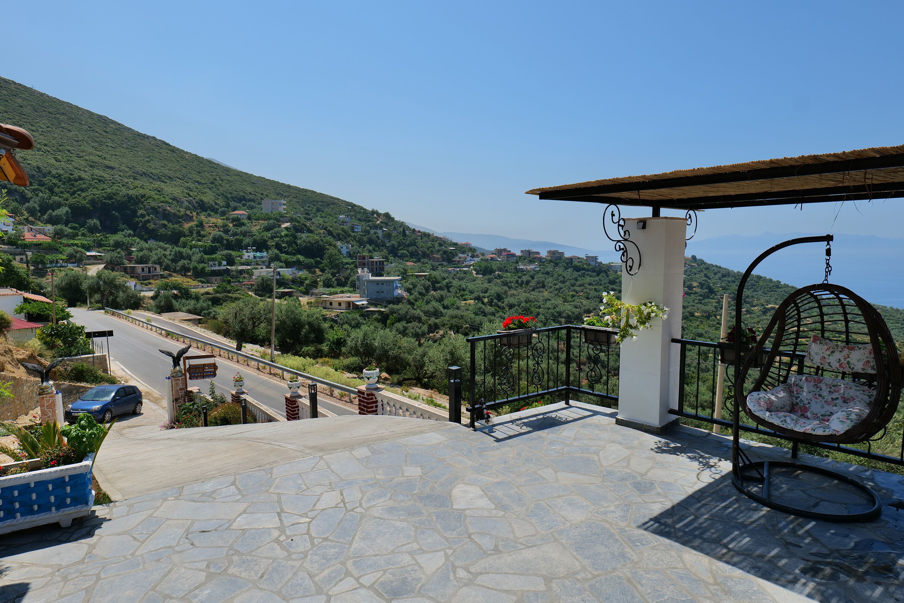Um terraço de pedra com uma cadeira suspensa, vista para uma aldeia no topo de uma colina e uma estrada descendo.