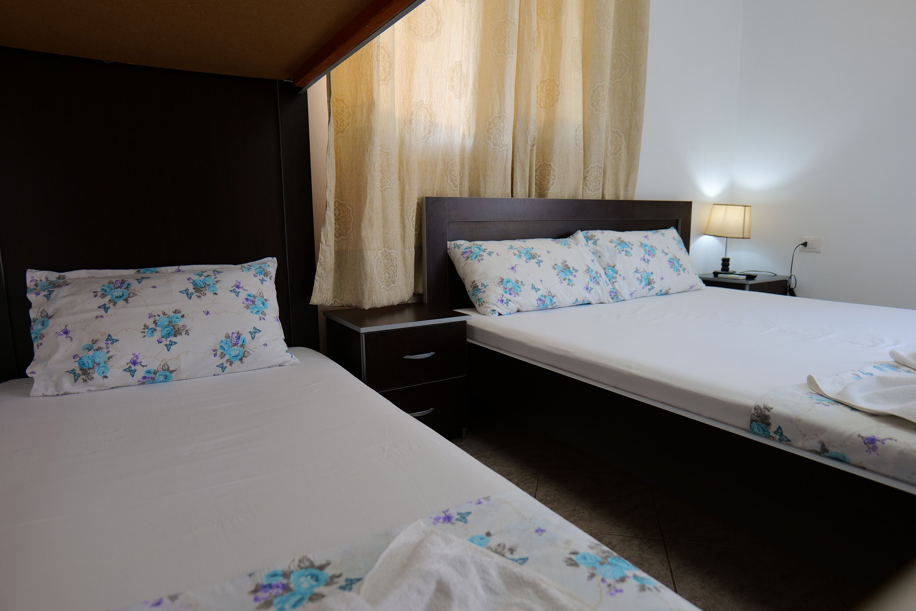 A hotel room with two beds, each with floral-patterned bedding, a nightstand with a lamp, and a window with curtains.
