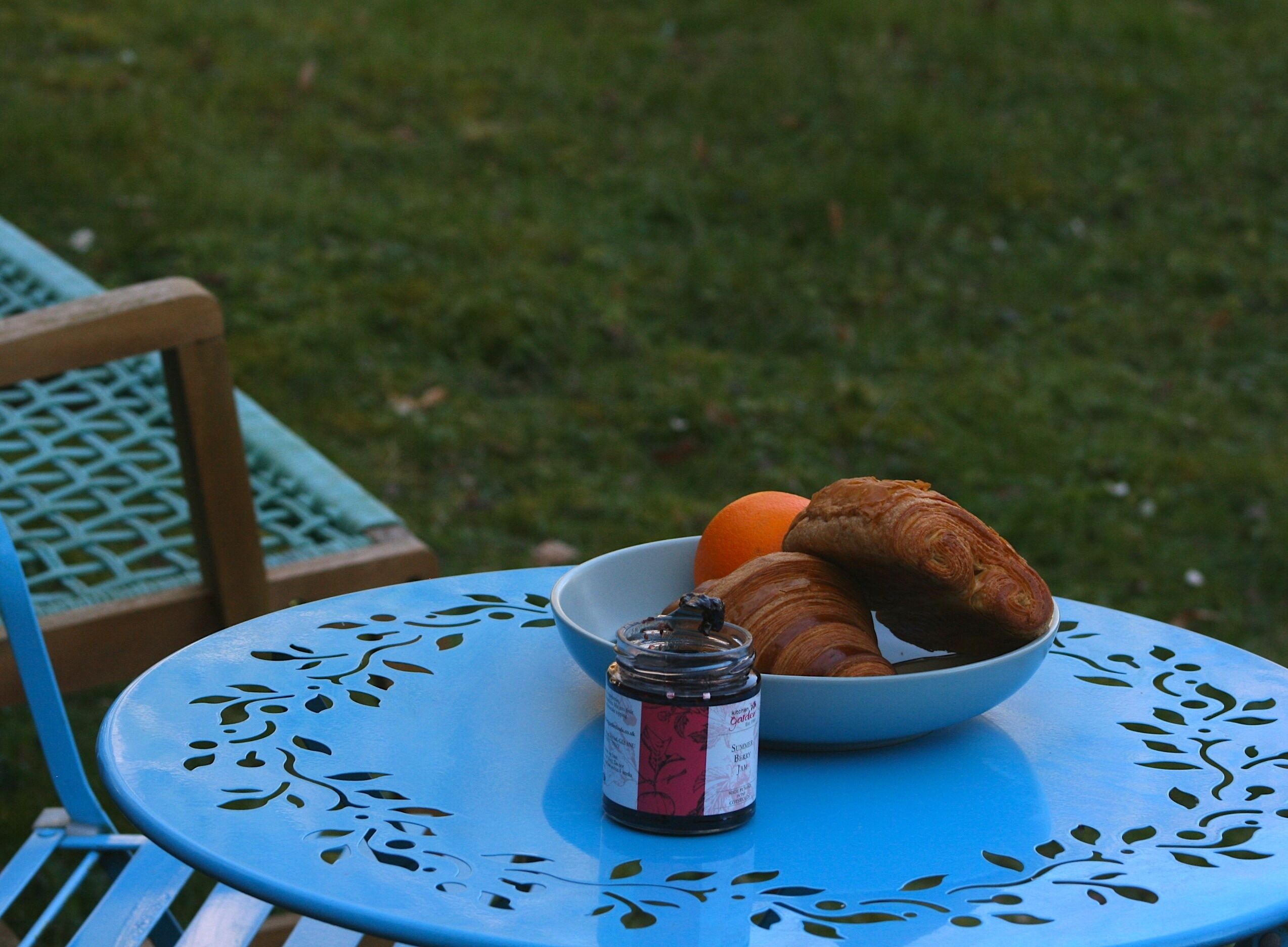 Ein runder Tisch mit floralem Muster, eine Schüssel Croissants, eine Orange und ein Marmeladenglas.