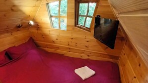 Een kleine, gezellige kamer met houten wanden en een schuine balkenlaag. Twee ramen zorgen voor natuurlijk licht, en aan de muur is een flatscreen televisie bevestigd. Het bed heeft een rode sprei met twee opgevouwen witte handdoeken.