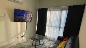 Un salon avec un canapé en cuir noir, une petite table, un ventilateur de plafond, un téléviseur fixé au mur et une fenêtre ornée de rideaux légers.