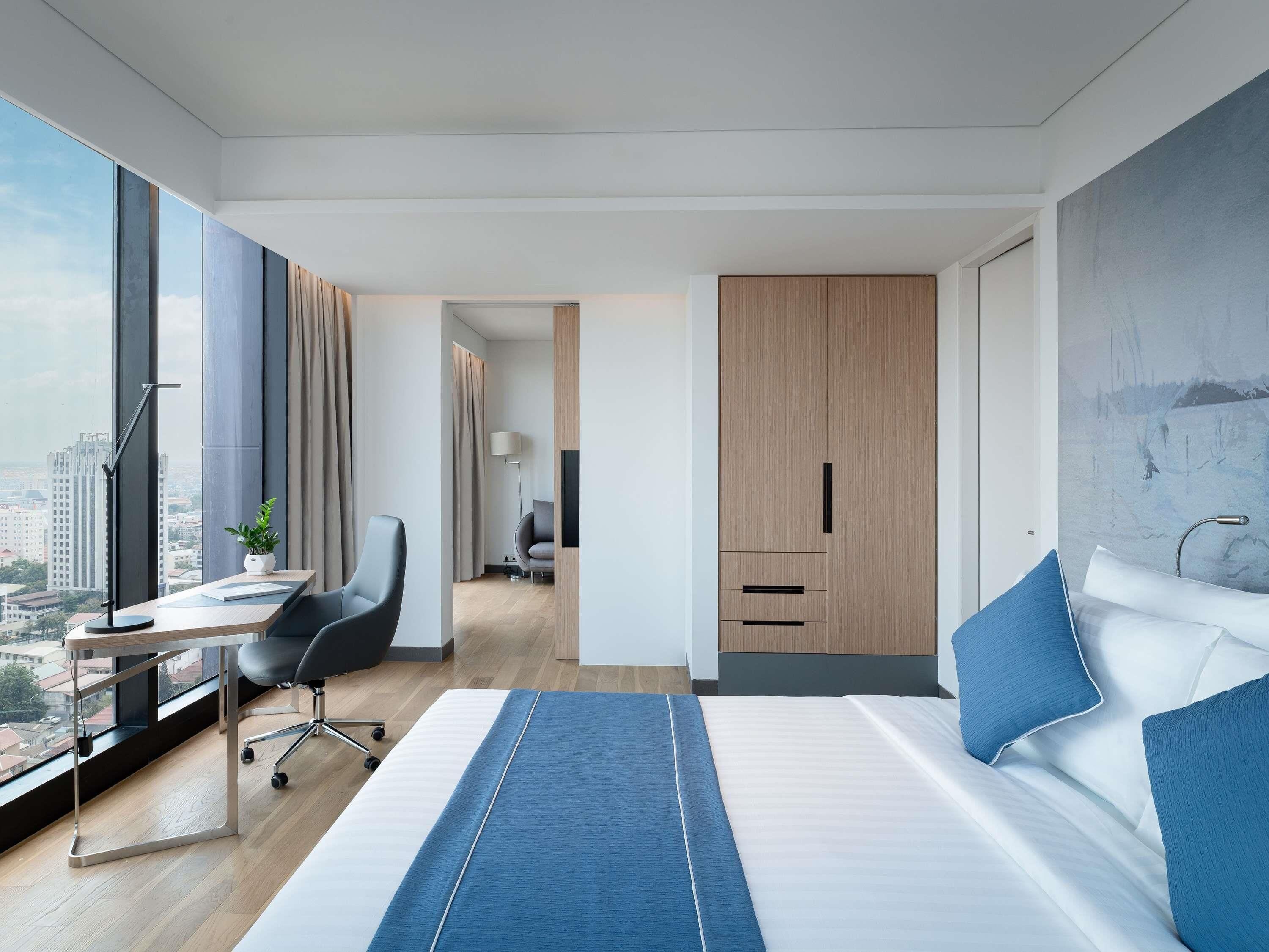 A modern hotel room with a large bed, a desk with a chair, a view of the city through large windows, and a wardrobe.