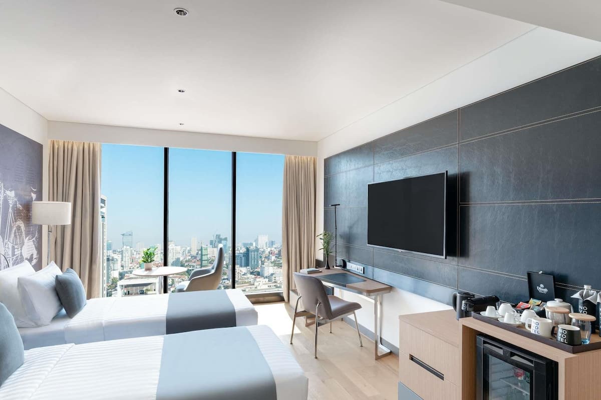 A modern hotel room with two beds, a desk, a coffee station, and a large TV.
