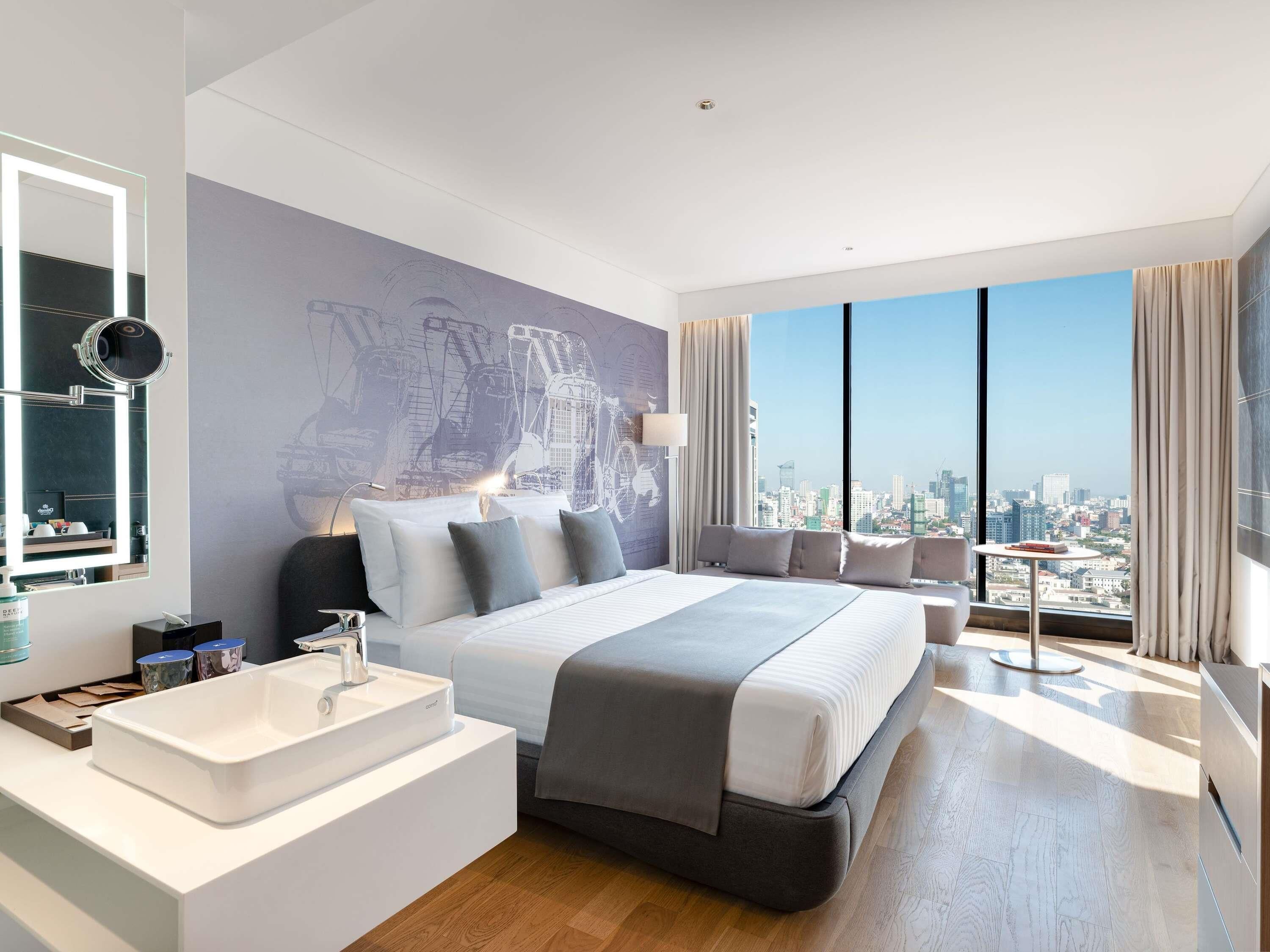 A modern hotel room with a large bed, a bathroom with a white sink, a city view, and a large window.