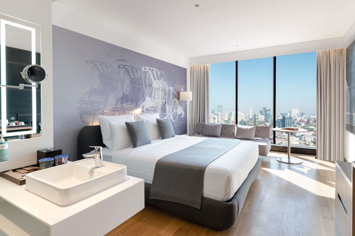 A modern hotel room with a large bed, a bathroom with a white sink, and a city view through large windows.