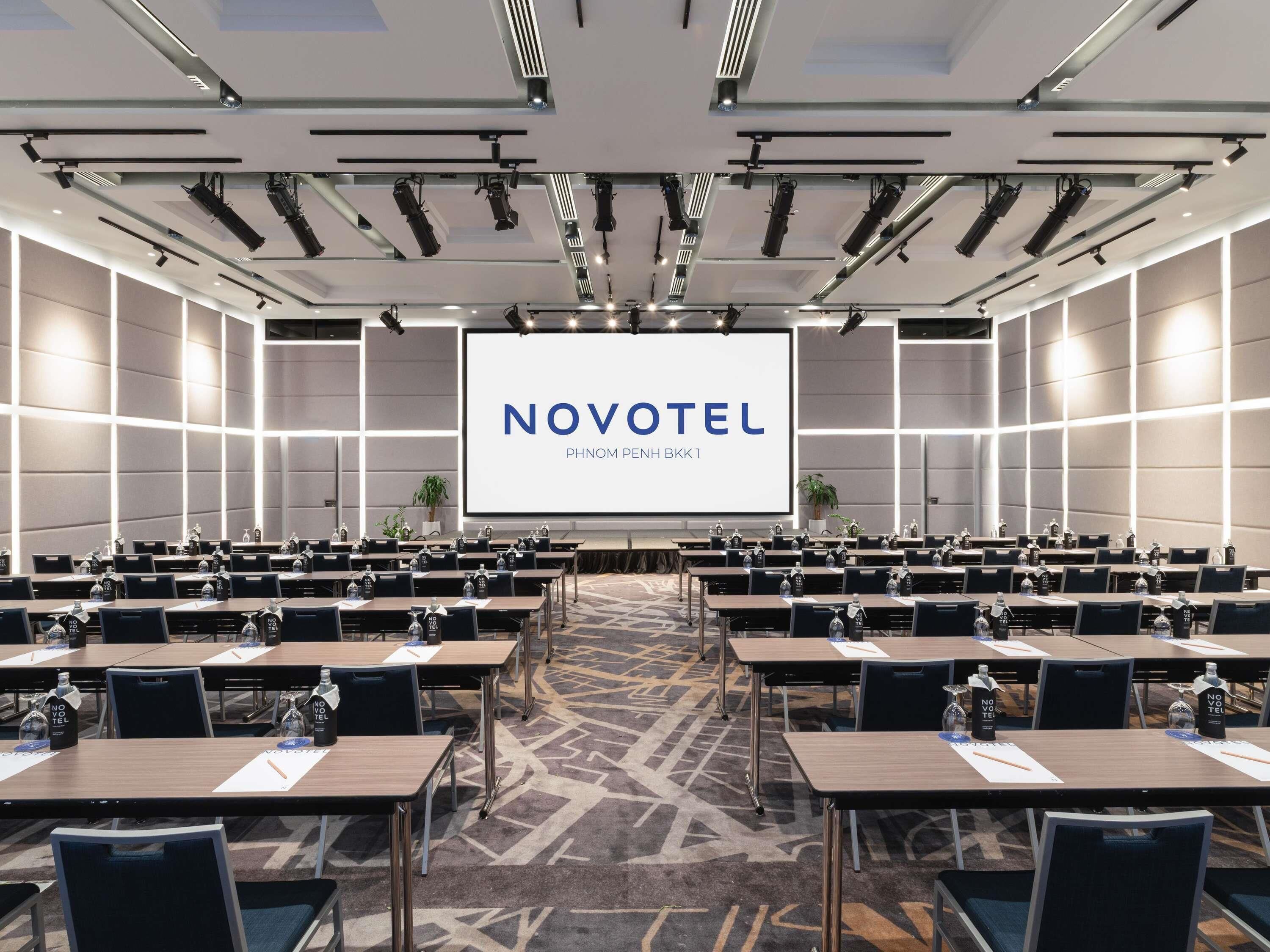 Une salle de conférence avec des rangées de tables, des chaises et un grand écran affichant le texte « NOVOTEL PHNOM PENH BKK1 ».
