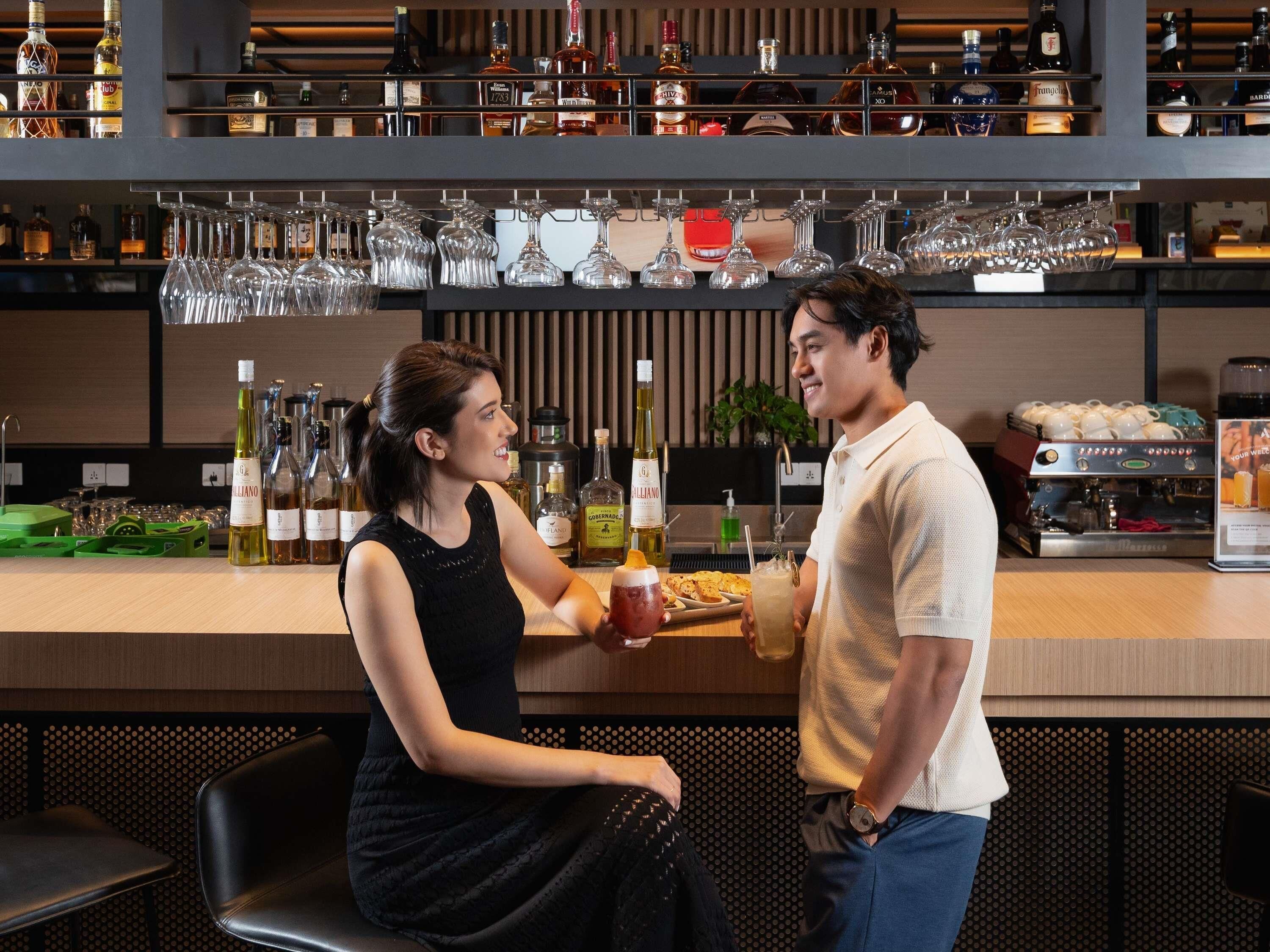 Uma área de bar com prateleiras de garrafas de bebidas e copos, uma máquina de café e um espaço de estar com cadeiras.