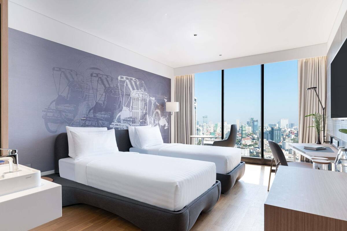Une chambre d’hôtel avec deux lits, un bureau, une chaise, une grande fenêtre donnant sur la ville et un mur orné d’un motif graphique.