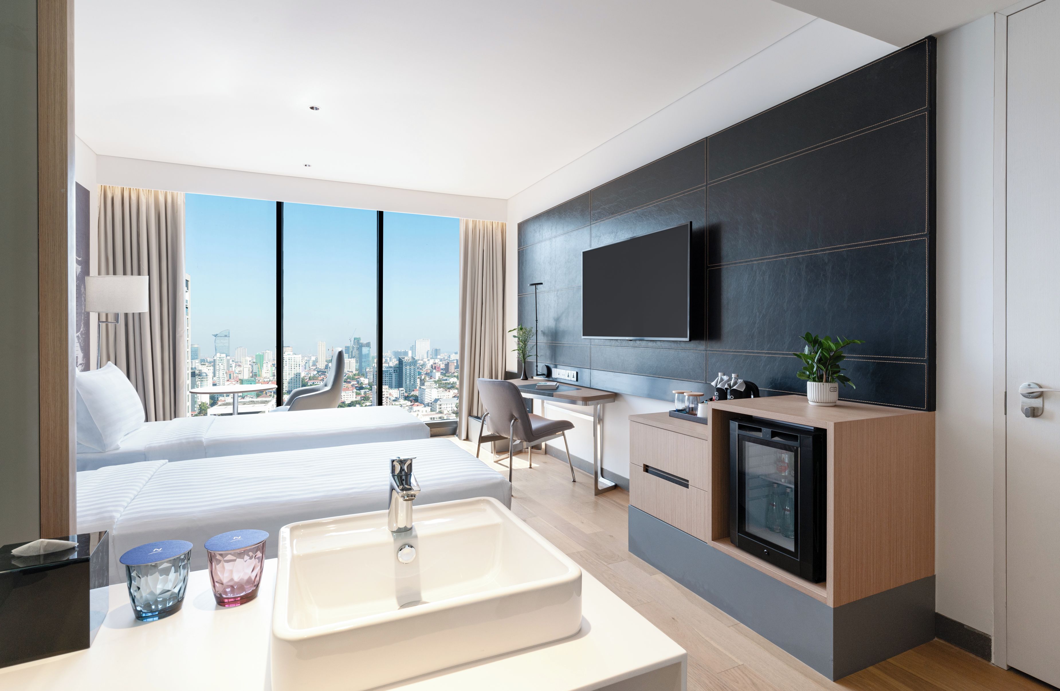 A modern hotel room with a city view, a bed, a desk, a TV, and a minibar.