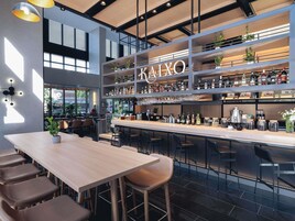 Un moderno bar con un letrero que dice 'KAIXO', taburetes, estantes con botellas y una zona de estar con mesas y sillas.