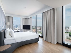 Ein modernes Hotelzimmer mit einem großen Bett, einem Flachbildfernseher, Blick auf die Stadt und einem großen Fenster mit Vorhängen.