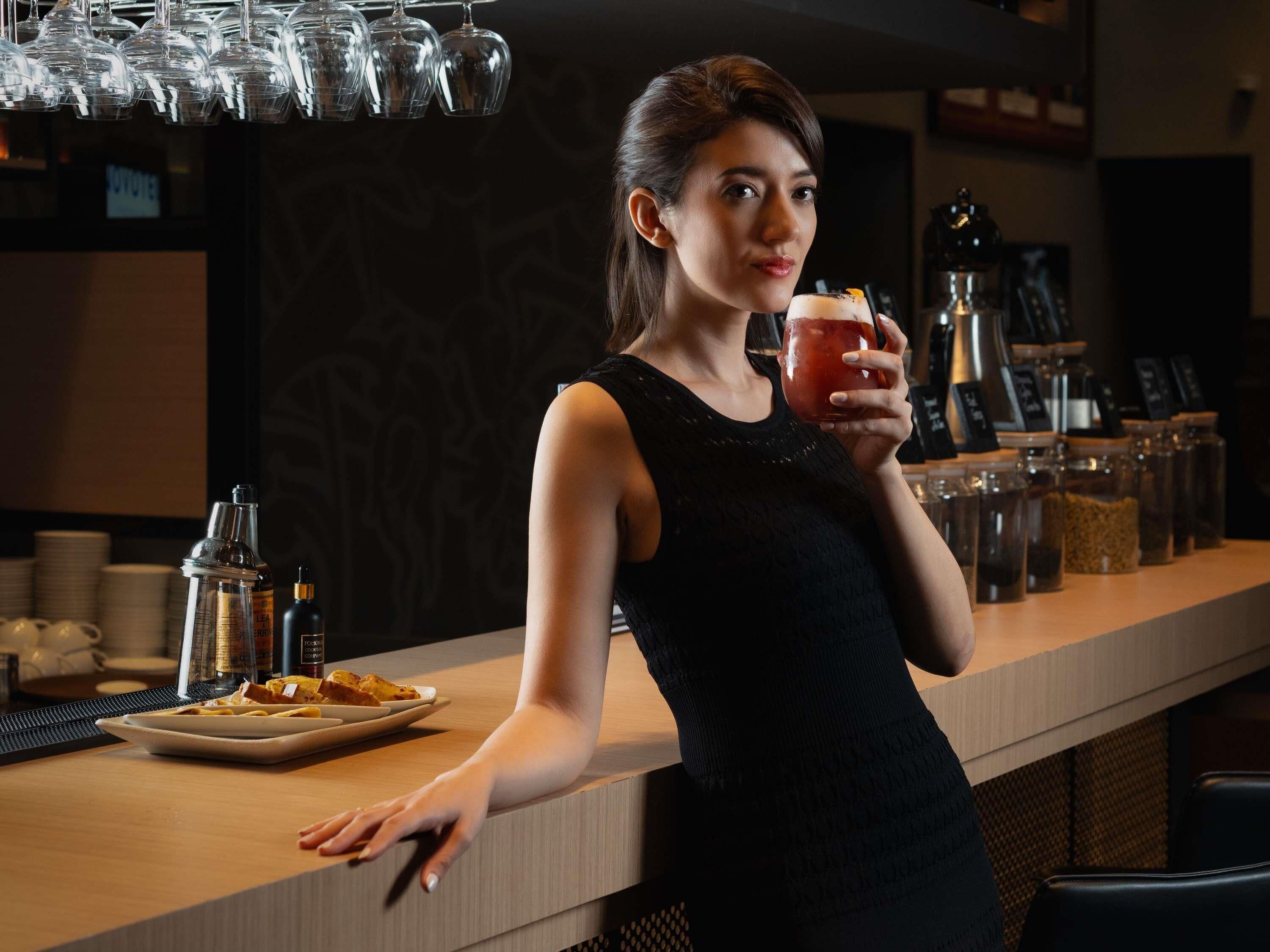 A person standing at a bar with a drink in hand, a plate of food, and various bar tools and ingredients in the background.