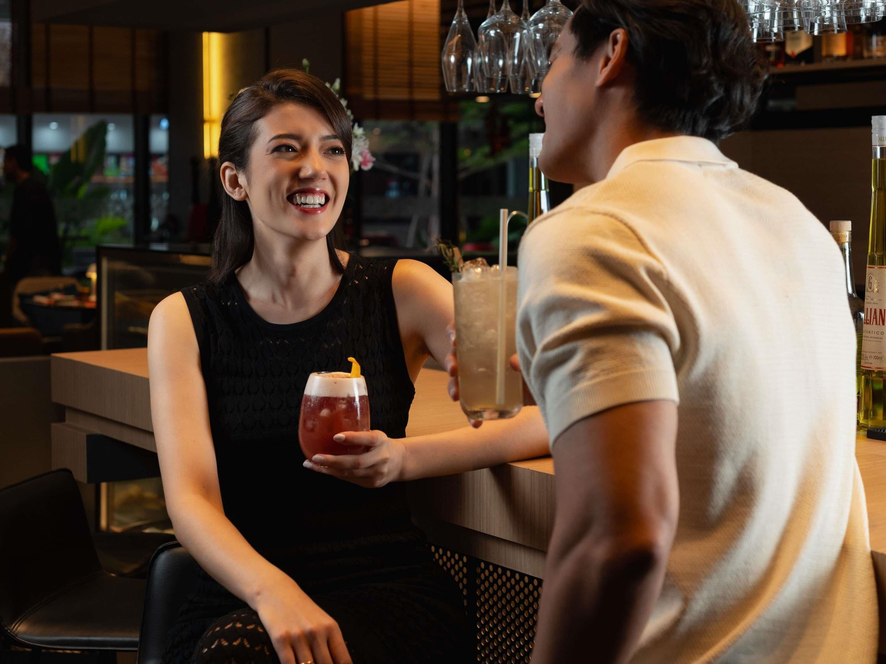 A person holding a drink with a straw, another person holding a glass, and a bar area with bottles in the background.