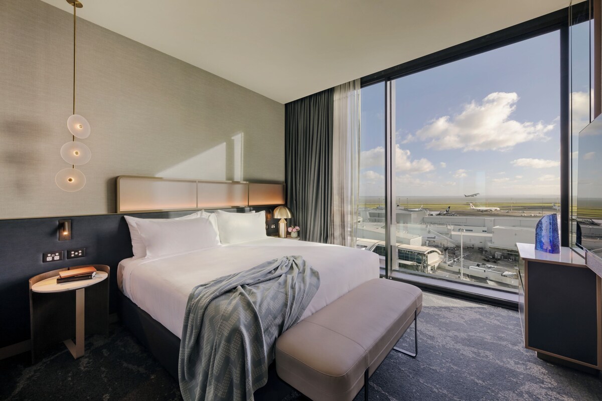 A hotel room with a large bed, a bedside table with a lamp, a bench, and a view of an airport.