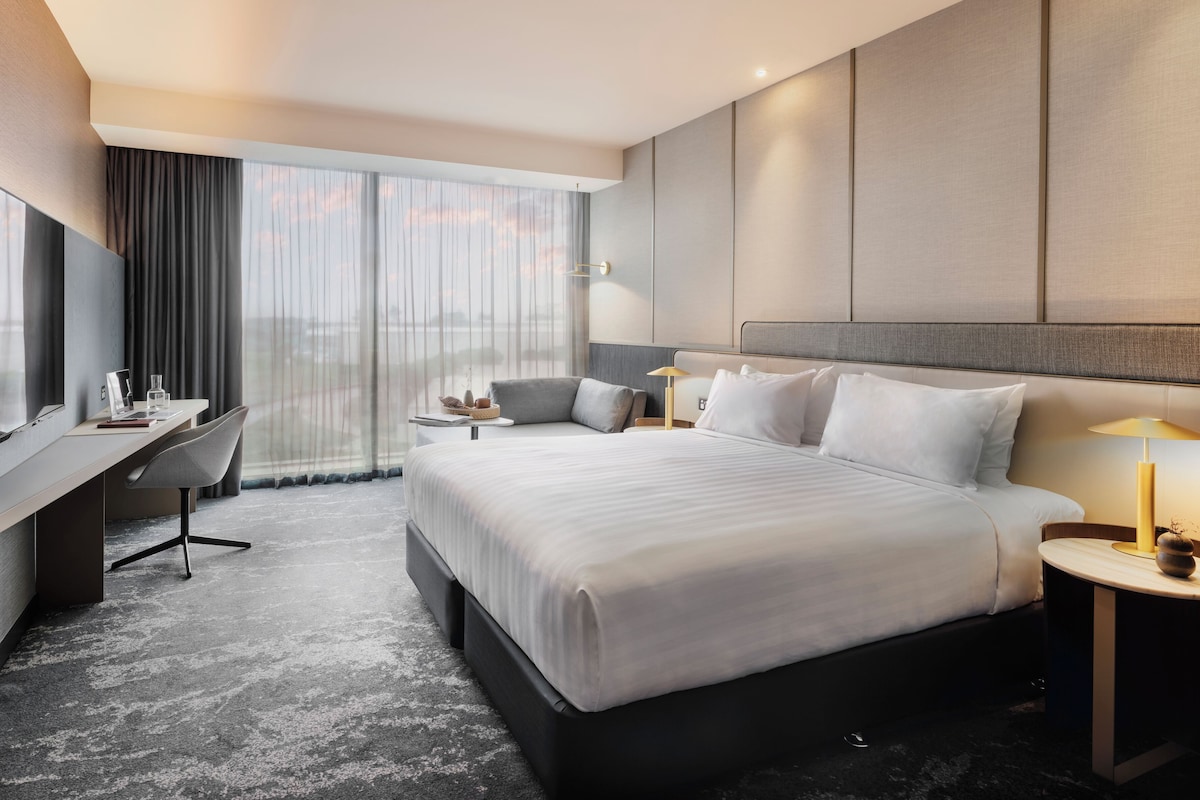 A hotel room with a large bed, a desk, a chair, a TV, and a window with curtains.