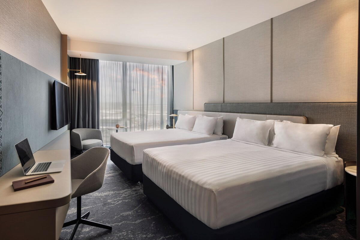 A hotel room with two beds, a desk with a laptop, a chair, a TV, and a window with curtains.