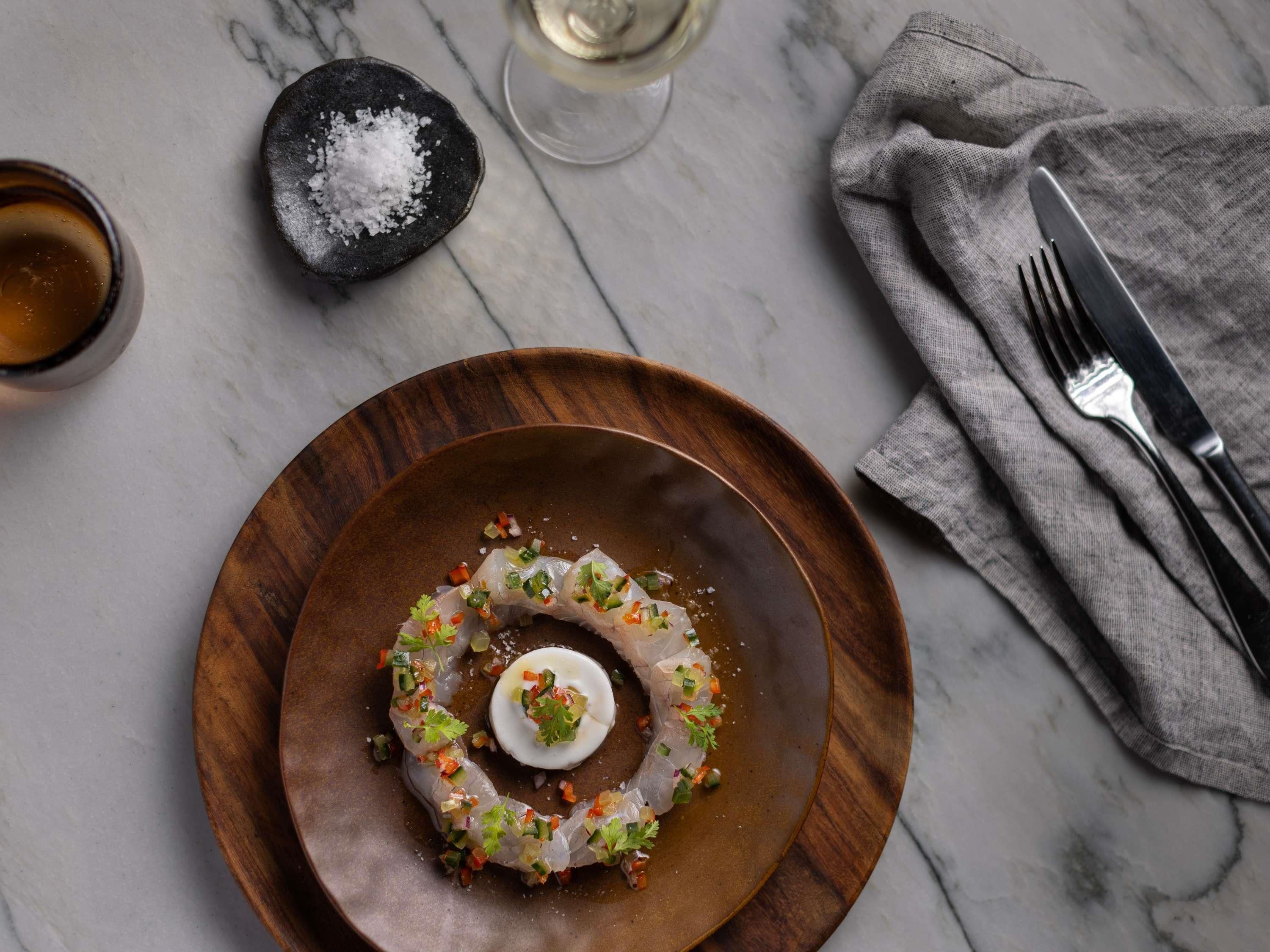 Un plato de madera con ceviche, una copa de vino blanco y un salero sobre una mesa de mármol.