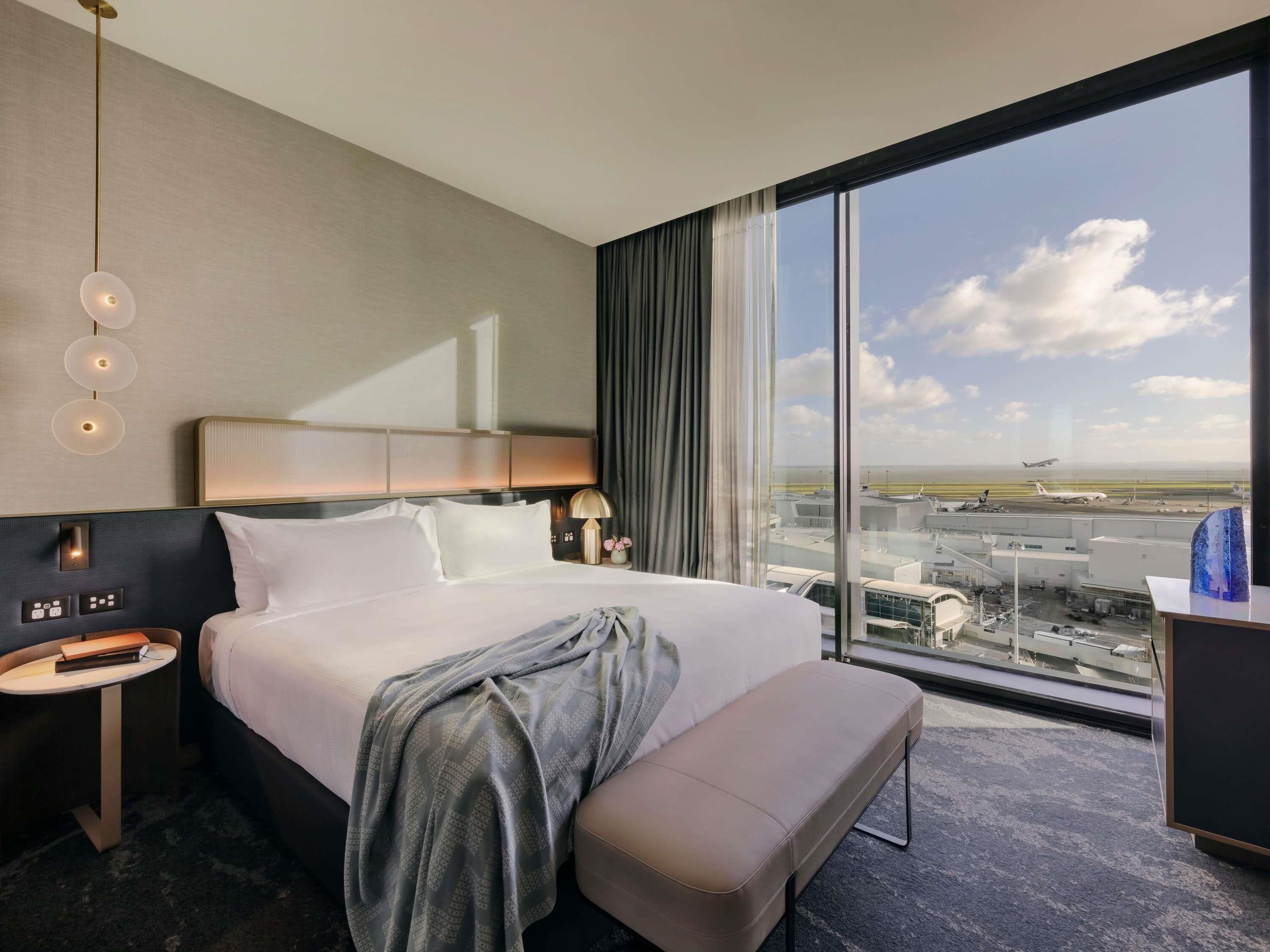 A hotel room with a large window, a bed, a bedside table, a lamp, and a bench.