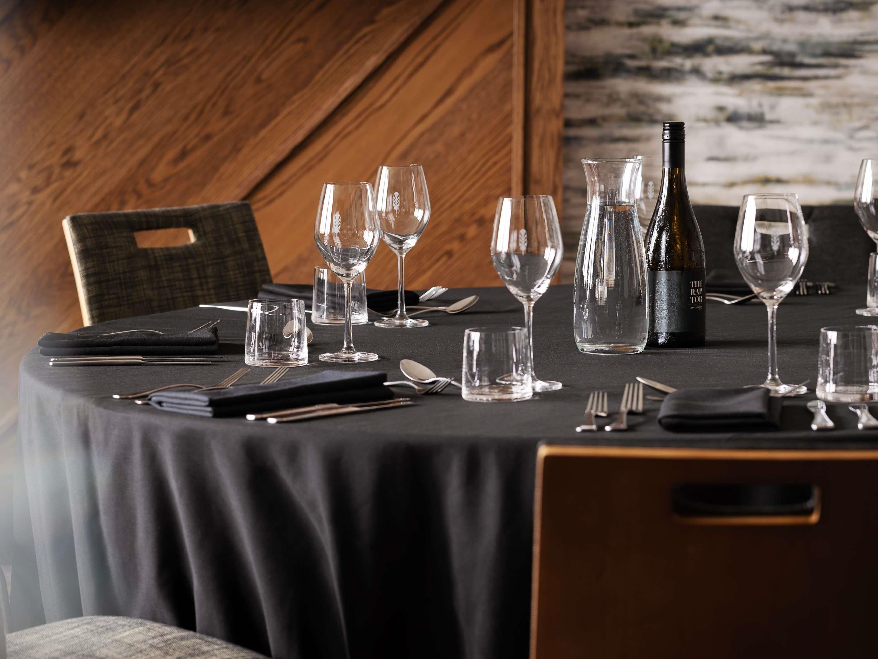 A dining table set with wine glasses, a bottle, and cutlery, with a wooden chair and a black tablecloth.