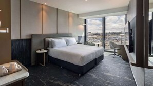 Moderni hotellihuone, jossa on suuri sänky, pieni yöpöytä, tuoli, televisio ja suuri ikkuna, josta avautuu näkymä kaupunkiin.