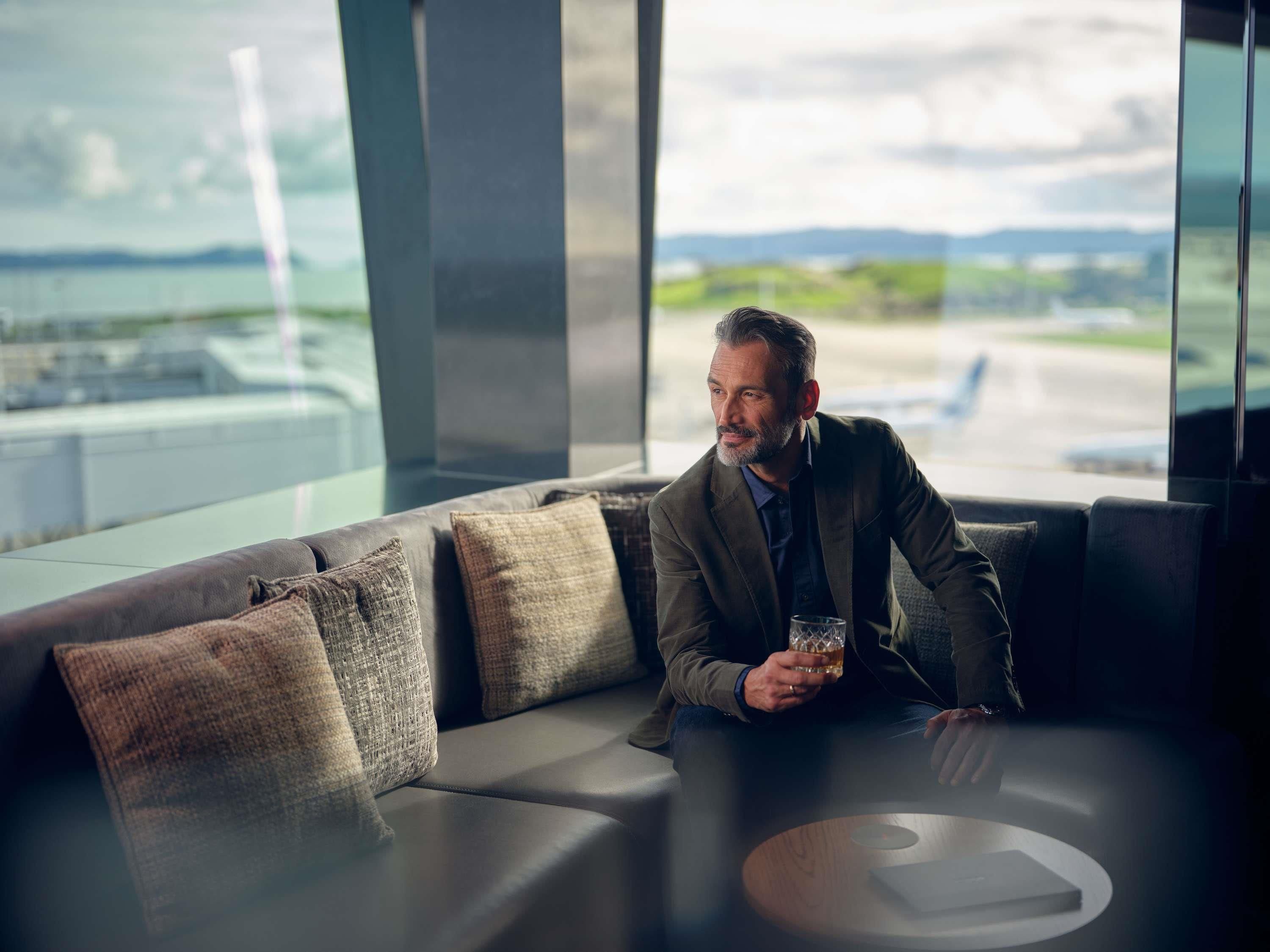 A man sitting on a modern sofa with a glass of whiskey, large windows with a view of an airport, and a minimalist design.