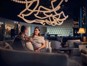 A couple seated on a couch in a modern lounge area with a geometric patterned wall and a decorative ceiling light fixture. - Pullman Auckland Airport (Auckland)