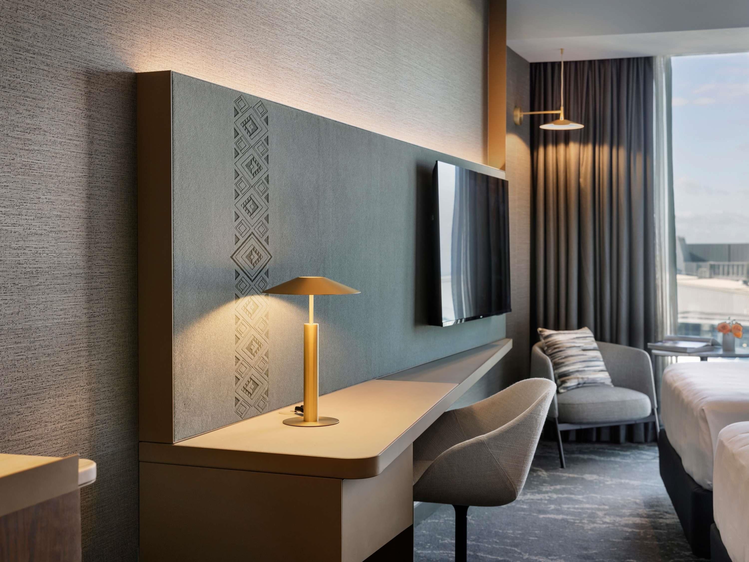 Habitación de hotel moderna con una gran televisión de pantalla plana en la pared, un escritorio con lámpara, una silla cómoda y una cama con vistas a la ciudad.